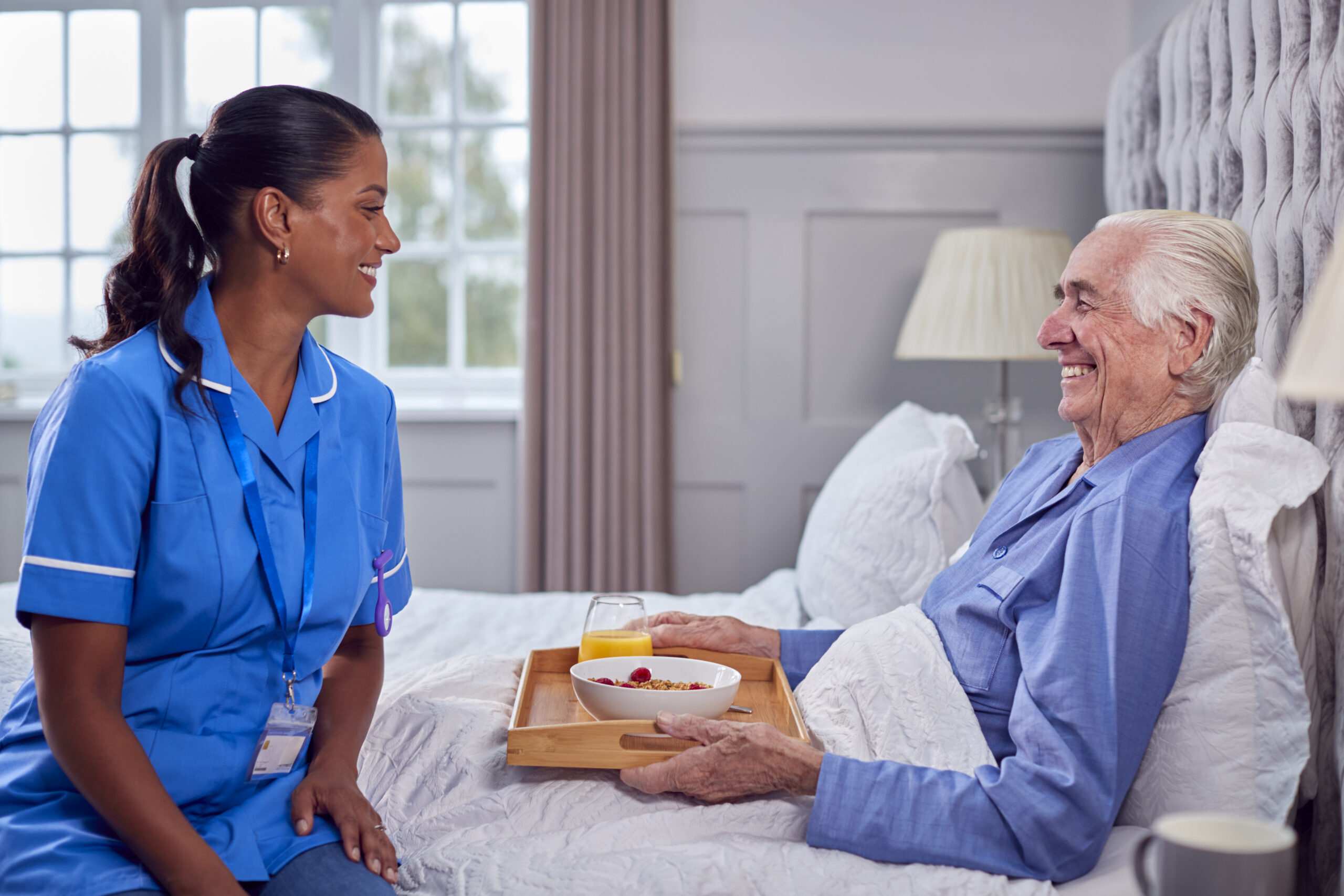 Female Care Worker In Uniform Bringing Senior Man At Home Breakfast In Bed On Tray